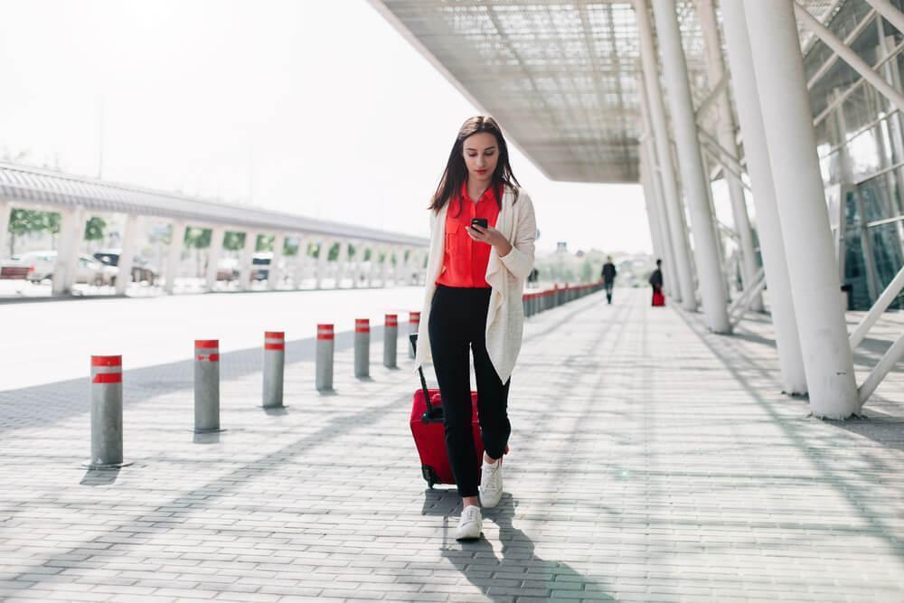 Woman walking in front of airport with bag and phone in hand